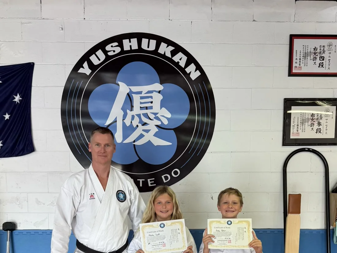 Sensei Sam Siegers with two students holding grading certificates at Yushukan Karate, dan certificates visible on the wall